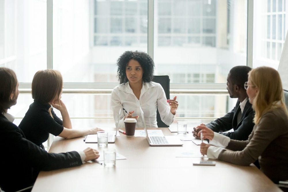 Woman of colour sitting at head of boardroom table talking 